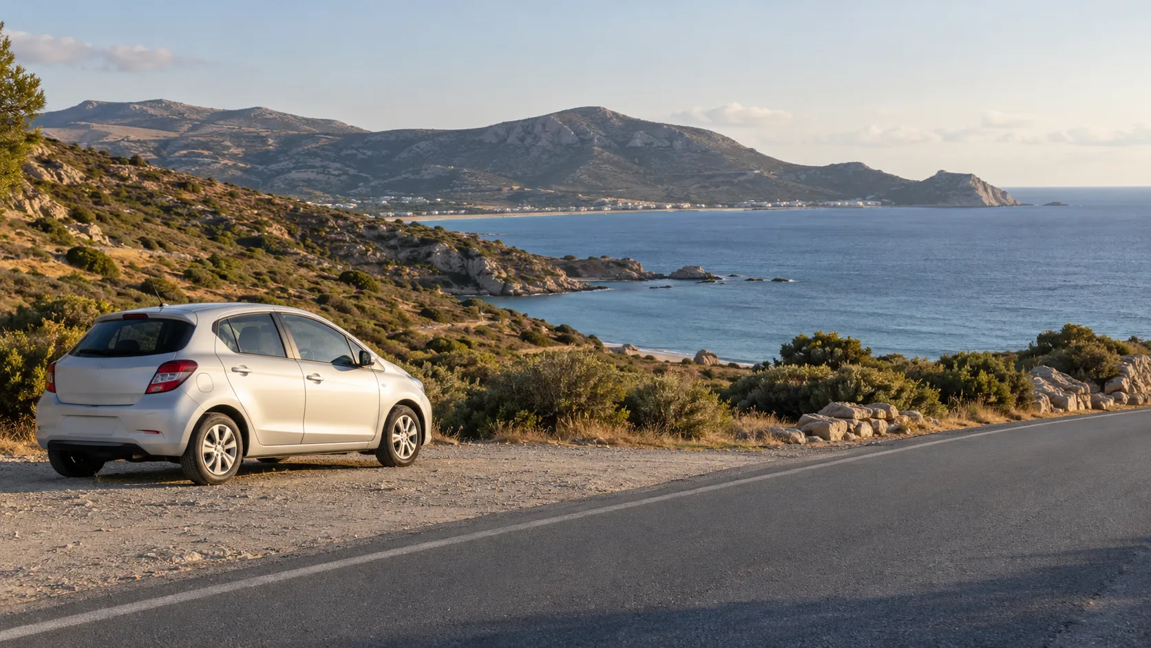 Compact car parked beside a coastal road with a wide bay and hills beyond